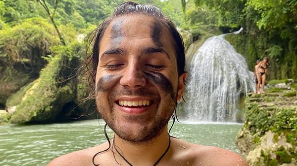 Deniz mit schwarzer Farbe im Gesicht, steht glücklich mit geschlassenen Augen vor einem Wasserfall
